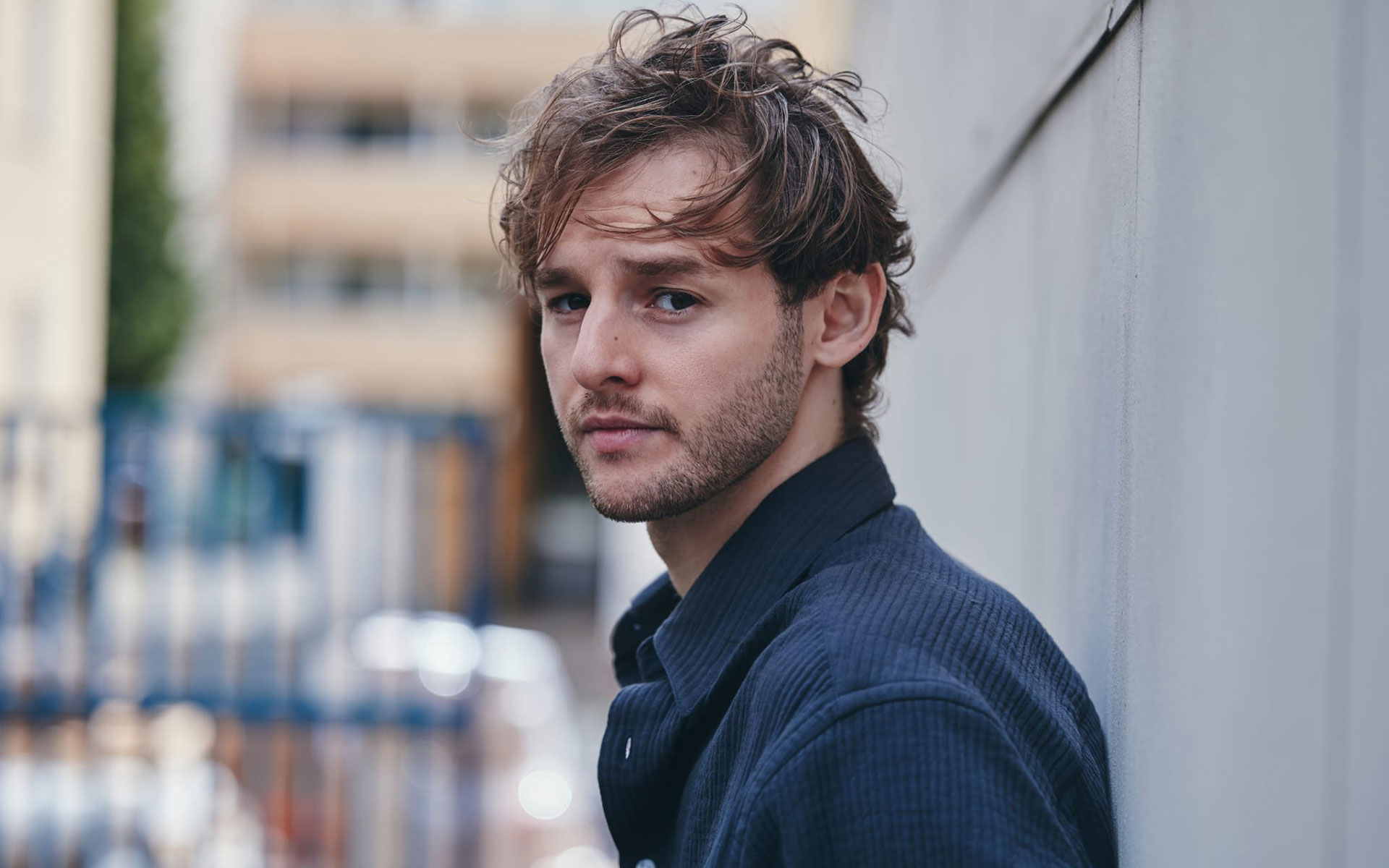Porträt eines jungen Mannes mit leicht zerzaustem Haar, der draußen an einer hellen Wand lehnt und ruhig in die Kamera blickt.